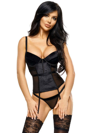 A woman with long dark hair is wearing the Beauty Night Bn6564 Monica Corset in black, featuring a corset-style top and matching panties with sheer tulle panels and lace detailing. She has garter straps attached to thigh-high stockings adorned with lace trim, and stands with her hands resting on her thighs.