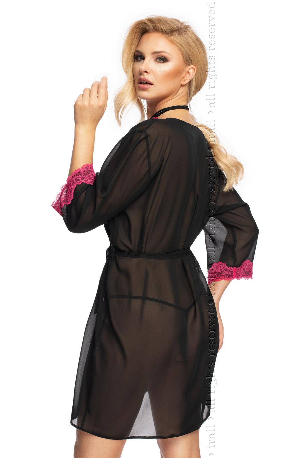 A woman with long blonde hair is posing in the Irall Erotic Flavia Dressing Gown, which is a sheer black chiffon robe featuring pink lace trim at the cuffs and a matching black belt. She is turned slightly to the side, looking over her shoulder with a serious expression. The gown is tied at the waist, and her hair is styled in loose waves.