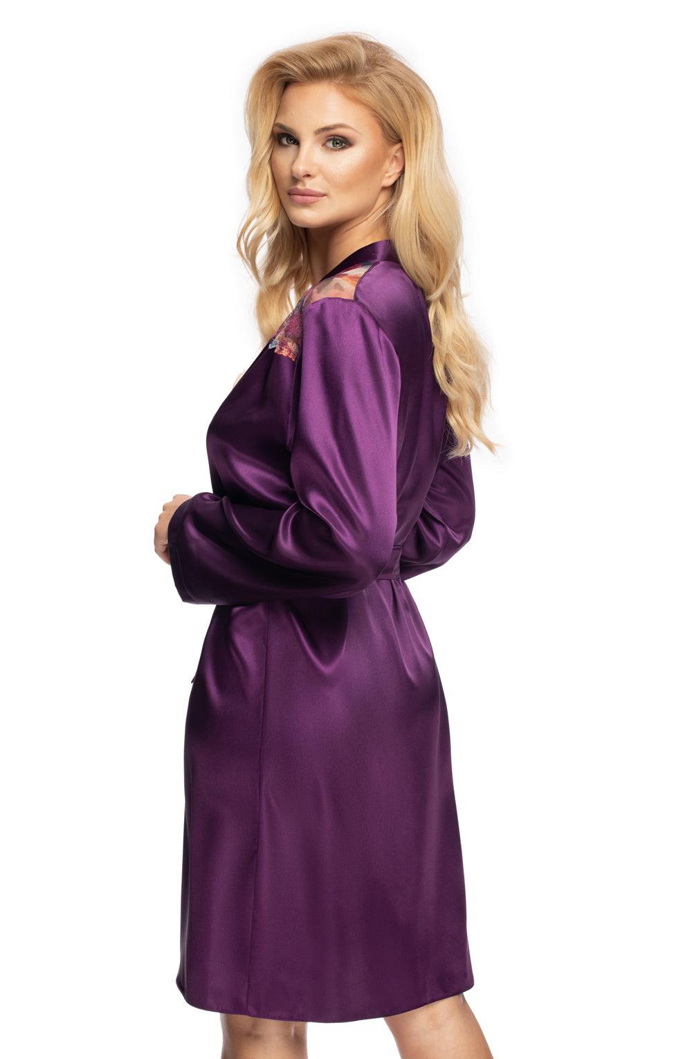 A woman with long, wavy blonde hair wears the Irall Shelby Dressing Gown Purple, featuring elegant lace details on the shoulders and a matching satin belt. She stands against a plain white background, looking over her shoulder with a subtle smile, her hands resting by her sides.