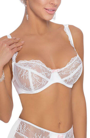 A woman is wearing a Roza Ambre Soft Cup Bra in white, featuring intricate floral lace patterns and a small pink bow in the center. She is also wearing matching lace underwear from the same collection. Her hand gently rests near her shoulder, adorned with dangling earrings. Her face is not fully visible.