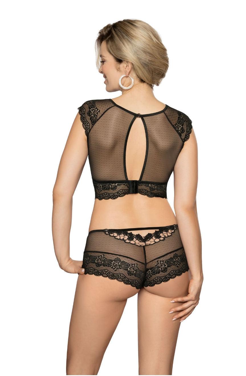 A blonde woman with short hair, part of the Roza Ellba collection, stands with her back to the camera, showcasing a black lace lingerie set. The Roza Ellba top features cap sleeves and a keyhole cutout, while the matching bottoms have intricate lace detailing. She accessorizes with large hoop earrings.