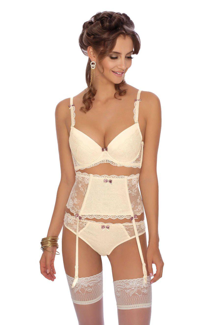 A woman is wearing an ivory-colored lingerie set featuring the Roza Fifi Push Up Bra, a high-waisted lace garter belt, panties, and thigh-high stockings. Her long, wavy hair is styled in loose curls and she accessorizes with gold earrings and bracelets. She looks down and smiles.