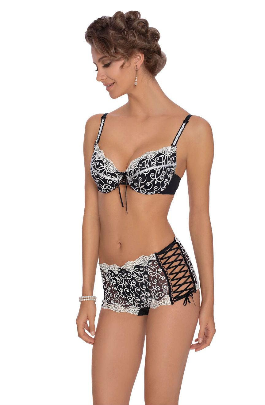 A woman is wearing the Roza Ginewra Bra, a matching black and white lingerie set from Roza. The bralette includes adjustable bra straps, lace trim, and a small bow in the center. The high-waisted bottoms feature lace embroidery and side laces. She has curly hair, pearl earrings, and a bracelet, smiling as she looks to her right.