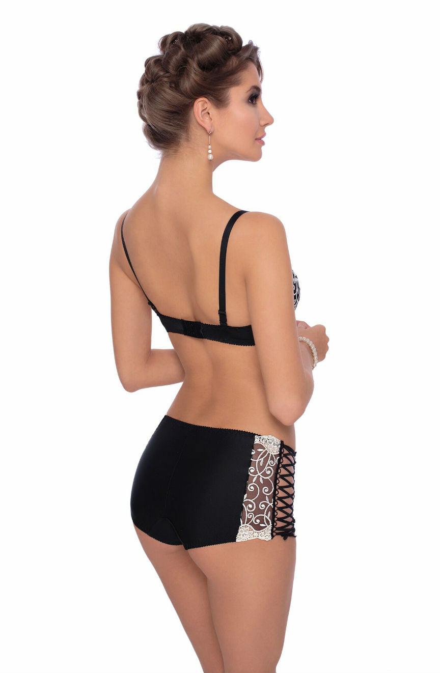 A woman is standing sideways, showcasing the back of her black Roza Ginewra lingerie set by Roza, which includes a bra with adjustable straps and high-waisted matching shorts featuring lace-up sides and floral embroidery. She has her hair styled in an updo and is wearing earrings and a bracelet. The background is white.