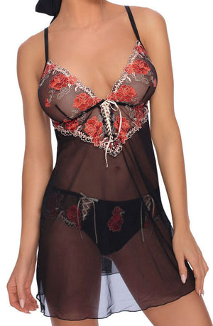 A person is wearing the Roza Natali Chemise Black from Roza, a sheer lingerie babydoll dress featuring red and white embroidered floral details on the chest area and matching sheer underwear. The dress has thin black straps and a lace-up front detail.