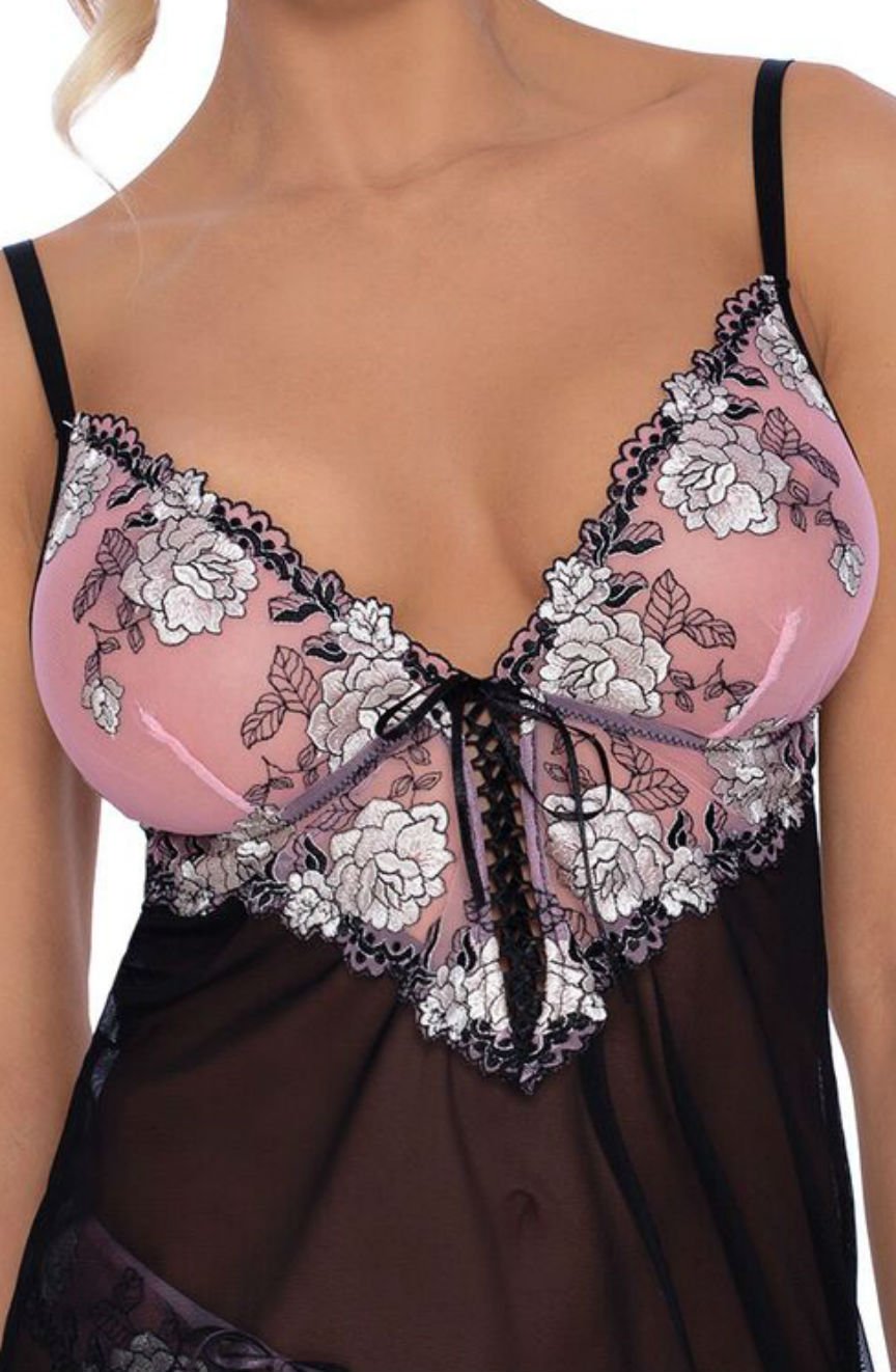 A close-up of a woman wearing the Roza Natali Chemise Pink from Roza. This delicate lingerie chemise features semi-transparent black fabric with a pink bust adorned with intricate white floral embroidery and black lace edging. Black thin adjustable straps and a central lace-up detail complete the design.