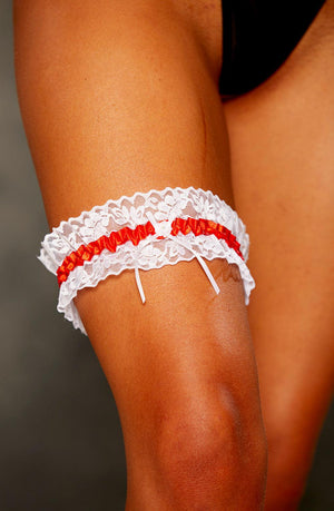 A close-up of a person's thigh wearing the Shirley Of Hollywood 19 Red Garter, a white lace garter with satin trim and a red ribbon from the brand Shirley of Hollywood. The person's skin tone is visible, and the background is blurred, highlighting the leg garter.